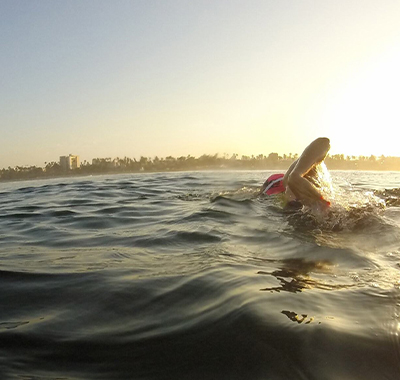 A woman swimming, with goggles and swim cap on, in open water with land in the background.  