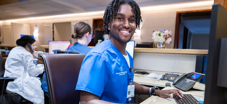 A male nurse in blue MSK scrubs sits at a computer and smiles. 
