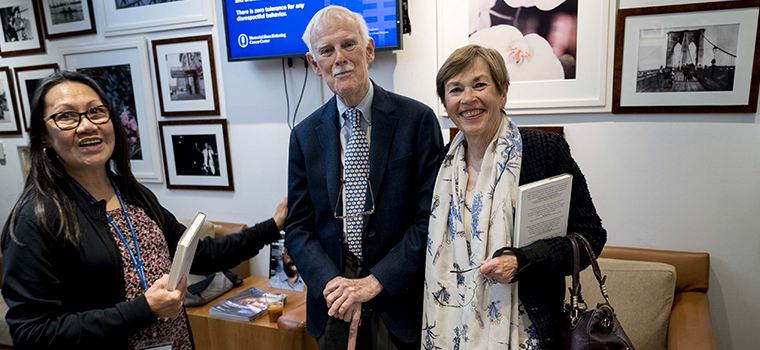 Three people standing in a photo-filled room, each holding a book, with a screen displaying a message in the background.
