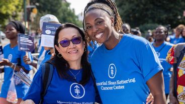 Two women wearing Memorial Sloan Kettering Cancer Center shirts smile, with their arms around one another. 