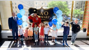 A group of children and Dr. Andrew Kung stand in front of an FAO Schwarz sign, smiling and saluting alongside a man dressed as a toy soldier. 