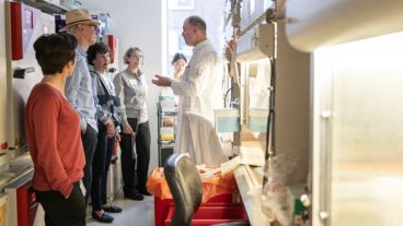 A group of elderly men and women stand, listening to an MSK scientist speaking inside a laboratory. 