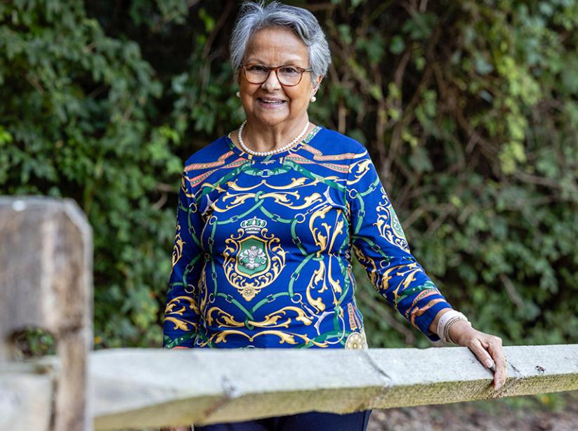 An older woman with short gray hair wearing a blue shirt with yellow, pink, and green designs stands with one hand on a wooden fence. 