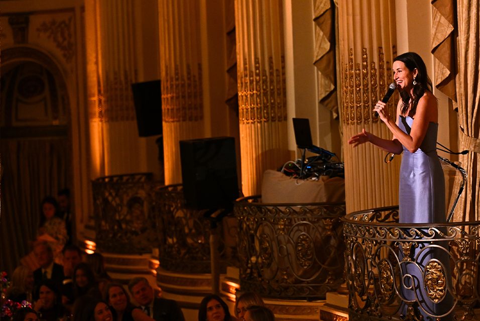 The event’s auctioneer, Lydia Fenet, stands at a podium holding a microphone.