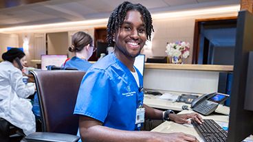 A male nurse in blue MSK scrubs sits at a computer and smiles. 