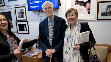 Three people standing in a photo-filled room, each holding a book, with a screen displaying a message in the background.