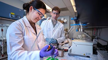 Two MSK scientists in a lab, both wearing lab coats and safety goggles.