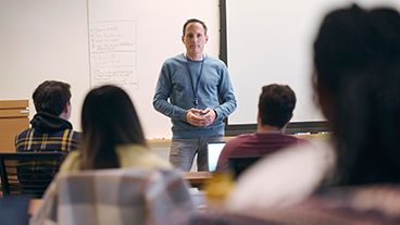 A teacher addresses a classroom with students seated in front of him.