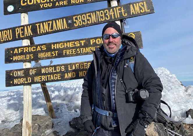 MSK donor John at Uhuru Peak in Tanzania. He is wearing a black parka and is standing in front of a sign that says "Africa's highest point."